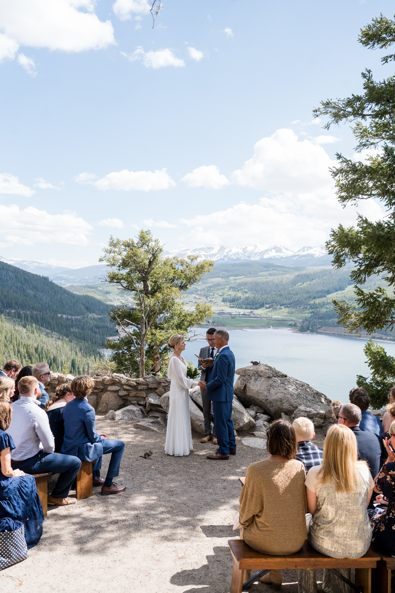 Bride and groom exchanging vows on a cliffside above Lake Dillon with wooden bench seating for guests